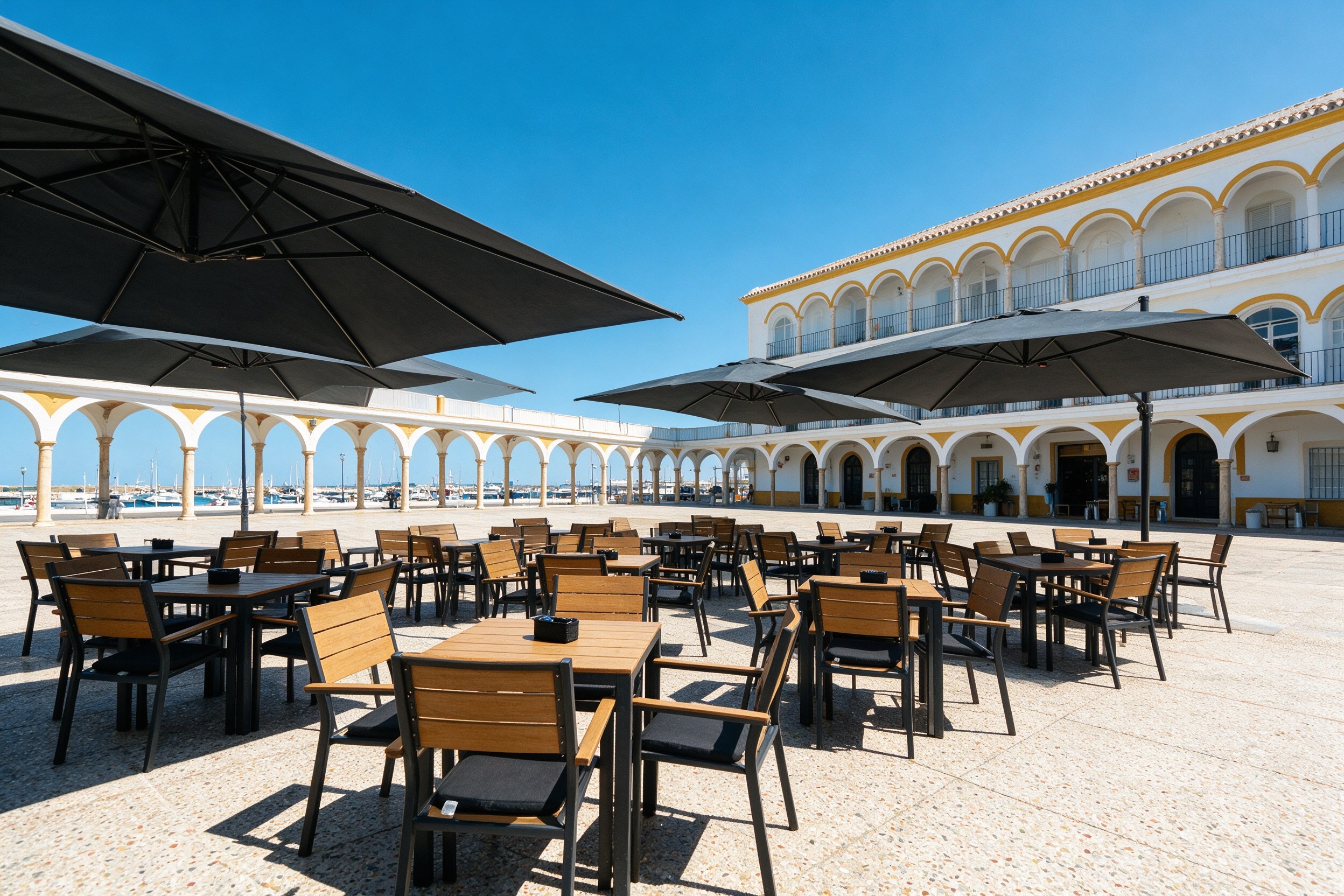Plaza portuaria con mesas y sombrillas de un restaurante bajo pórticos amarillos y blancos